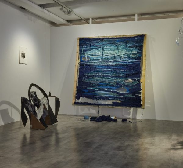 A gallery installation features a large textured artwork in shades of blue mounted on a white wall, with pieces of fabric on the floor beneath it. To the left is a dark abstract sculpture, and to the right, a hanging mobile made of curved metallic and black elements.