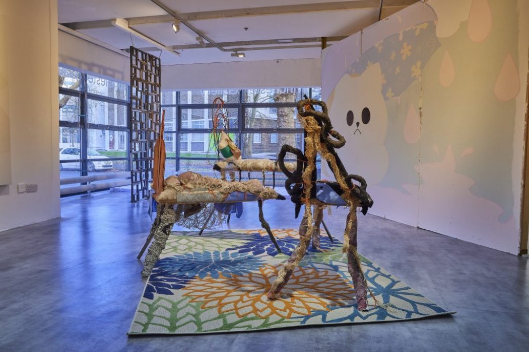A contemporary gallery installation features abstract sculptural pieces made of textured materials arranged on a colorful floral-patterned rug. The background includes large windows and a wall mural with soft pastel shapes and a cartoon-like face.