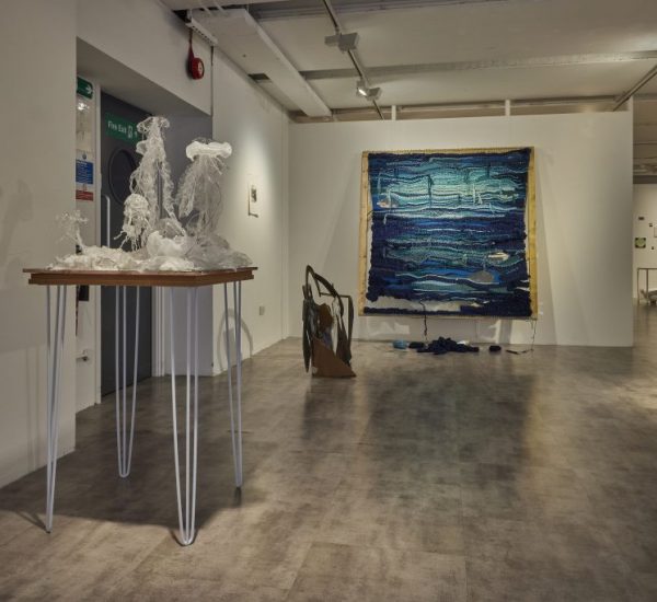 An art gallery with a white-walled interior displays various contemporary artworks, including a translucent sculpture on a tall table, a large blue textile piece on the back wall, and hanging abstract installations. The floor is gray and the space is lit with ceiling spotlights.