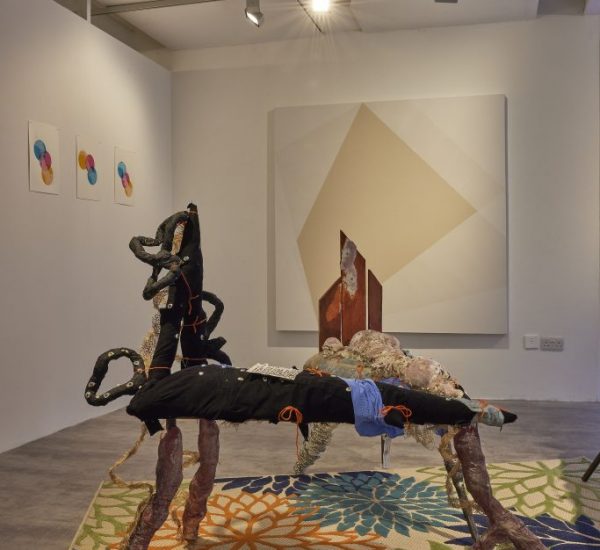A gallery installation features two abstract sculptural pieces made of mixed materials positioned on a colorful floral-patterned rug. Behind them, a large geometric painting hangs on the wall, with three smaller circular artworks to the left and a window to the right.