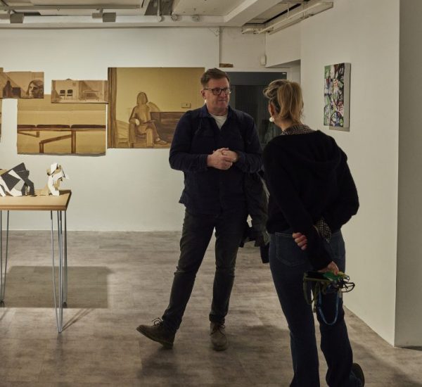 Two people stand in an art gallery near a wooden table displaying abstract black and white sculptures, with several sepia-toned portraits and interior scenes mounted on the wall behind them. Another colorful artwork is visible on the adjacent wall, and part of a third person is seen on the right edge of the image.