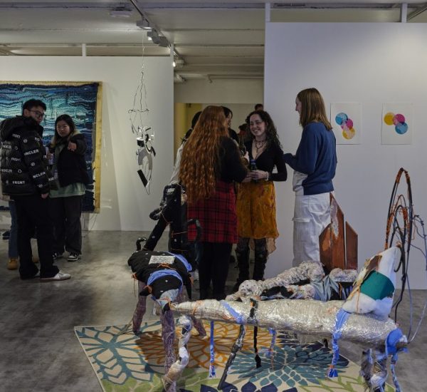A group of people stand and talk in a contemporary art gallery featuring colorful abstract artworks on the walls and mixed-media sculptures on the floor. The space includes a large blue textile piece, hanging installation, and a patterned rug with sculptural elements.