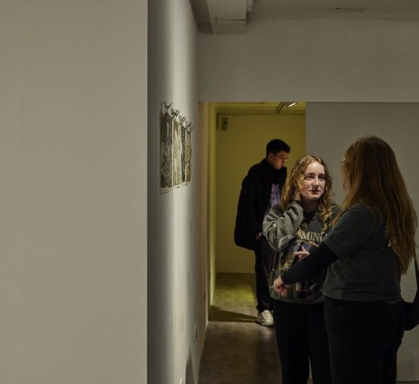 Two people stand in a narrow gallery hallway looking at small artworks displayed on the wall, while another person is visible further back in the corridor. A single framed piece hangs on the left wall, and the space is lit with overhead lighting.