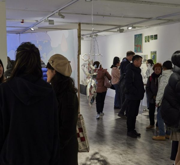A group of people are gathered in a contemporary art gallery, viewing and discussing artworks displayed on white walls. The space features hanging sculptures, framed pieces, and colorful paintings arranged throughout the room.