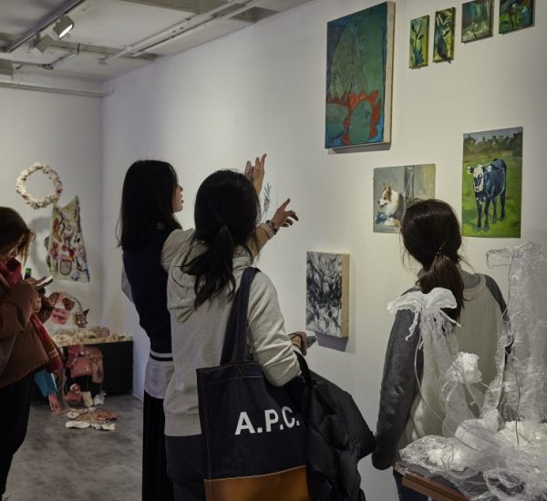 Several people are viewing and discussing artworks displayed on a white gallery wall, which includes paintings of landscapes, animals, and abstract designs. Sculptural pieces made of translucent material and other mixed-media works are arranged on tables and the floor nearby.