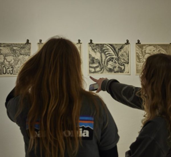 Two people stand in front of a white wall examining four monochrome drawings clipped in a row. One person is pointing at a drawing while holding a phone, and the artworks feature abstract and organic shapes.