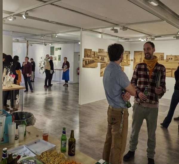 A gallery space with white walls displays architectural drawings and models, while several people stand and converse in small groups. In the foreground, a table holds drinks, snacks, and empty cups, suggesting a social event or exhibition opening.