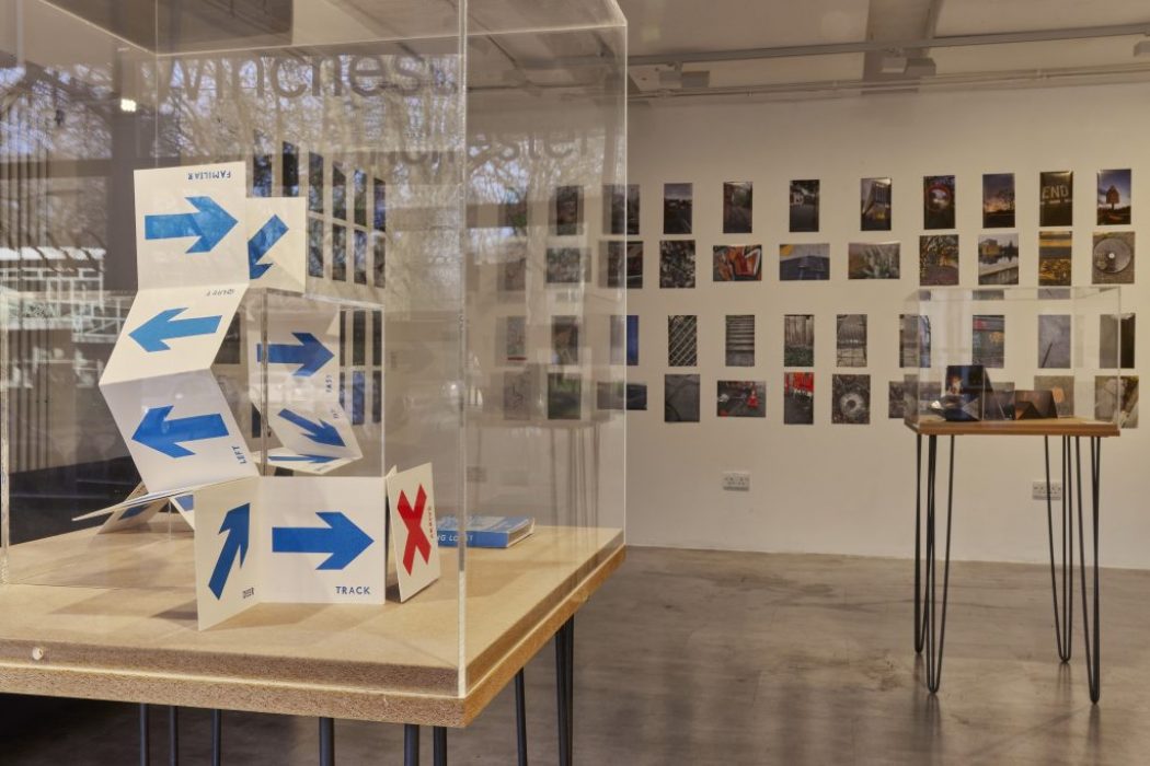 An art gallery display features a folded paper structure marked with blue arrows and a red 'X' inside a glass case. Behind it, a wall is covered with small photographs arranged in a grid, and another table with items is visible to the right.