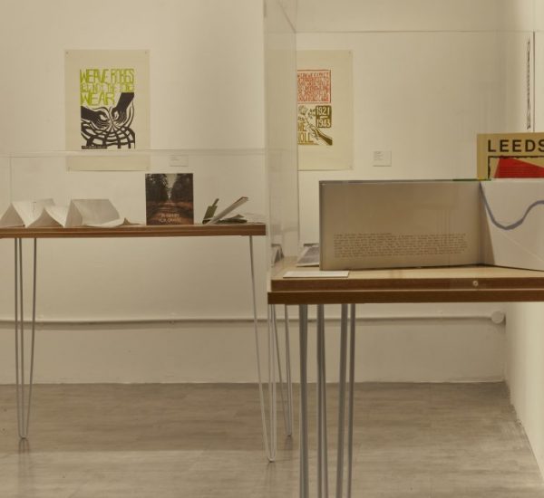 An art exhibition features two display tables with books and paper models enclosed in glass cases. Framed posters hang on the walls behind, including one with the word "LEEDS" in bold letters.