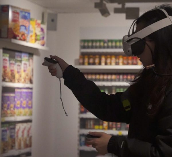 A person wearing a virtual reality headset and holding controllers interacts with a simulated environment in a room designed to look like a grocery store aisle. Shelves in the background are stocked with colorful cereal boxes and jarred goods.