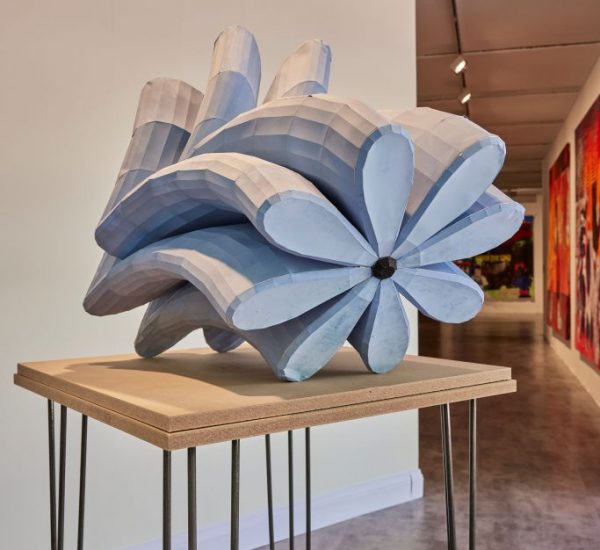 A light blue geometric sculpture with petal-like shapes is placed on a wooden table with metal legs in a gallery setting.