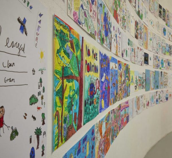 A close-up view of a curved wall shows vibrant children’s drawings, including messages such as “No More Pollution” and nature scenes. The display emphasizes creativity and environmental awareness.