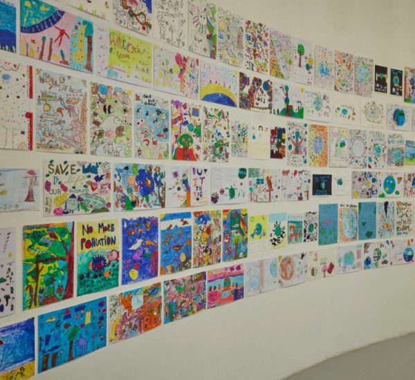 A curved gallery wall is covered with colorful drawings and posters created by children, featuring themes like nature and environmental messages. The artworks are arranged in neat rows across the white surface.
