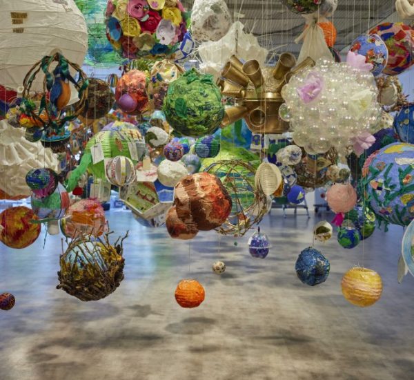 An art installation features numerous colorful spherical objects suspended from the ceiling by thin strings. The spheres vary in size and design, creating a vibrant floating display in a spacious gallery.