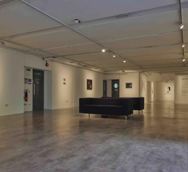 A wide view of the gallery shows multiple paintings on white walls, including a yellow canvas with a faint bicycle outline and vibrant abstract pieces. Black seating is arranged in the center of the room under track lighting.