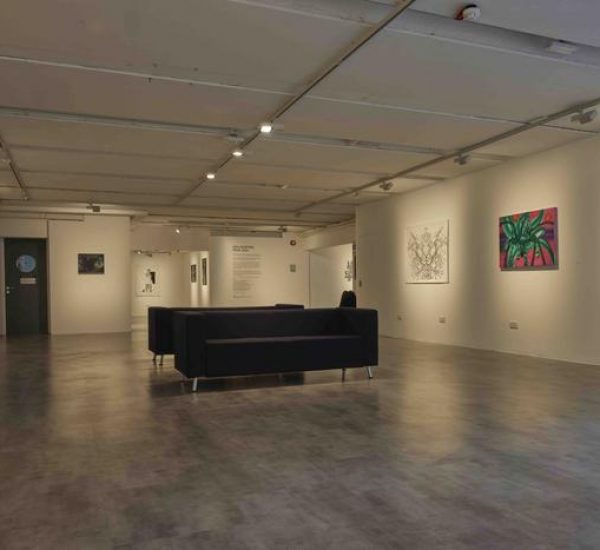 A spacious gallery interior features artworks on both walls, including a green palm-like painting and intricate black line drawings. Two black sofas sit in the center, creating a viewing area under bright ceiling lights.