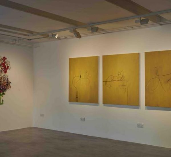 Three yellow-toned paintings with minimalist line drawings of bicycles and animals are mounted on a wall next to a colorful abstract artwork. The gallery floor is gray and the lighting highlights each piece.