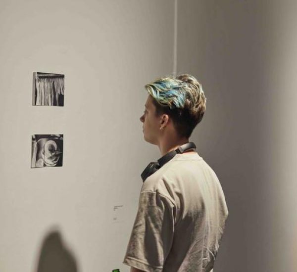 A person observes two small monochrome artworks on a white wall, with a dark abstract painting visible in the background.