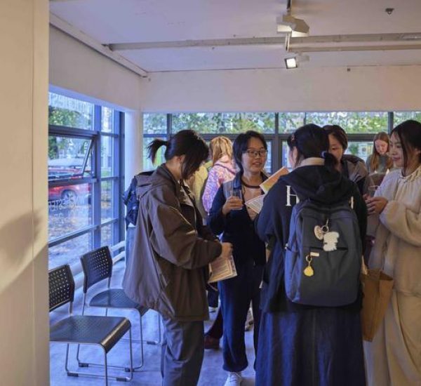 Several people stand near a windowed area of the gallery, holding papers and drinks while conversing.