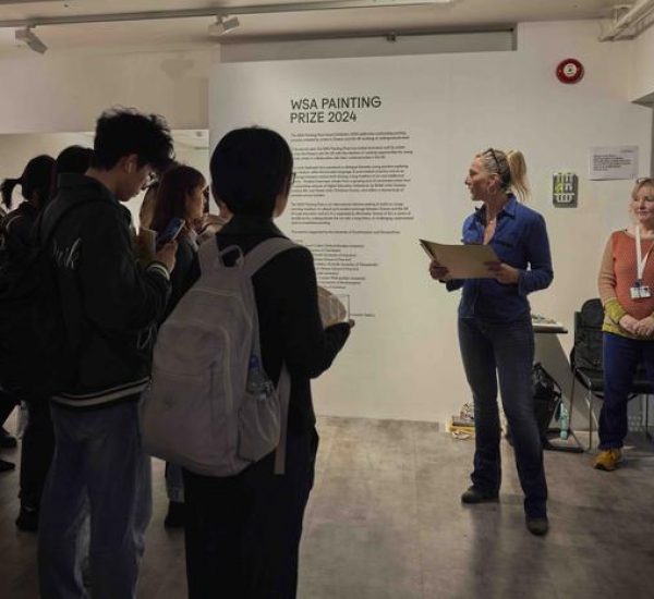 A person speaks to a group in front of a wall with “WSA Painting Prize 2024” text, while others listen and hold papers in the gallery setting.