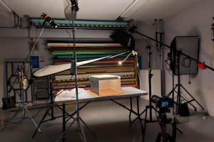 A professional photography studio setup featuring lighting equipment, a camera on a tripod, and a small object on a table being photographed against colorful backdrop rolls.