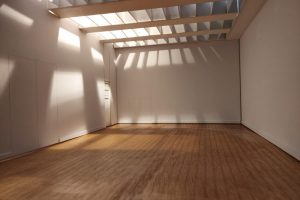 An empty room with white walls, wooden flooring, and a beamed ceiling that casts sunlight patterns across the space.