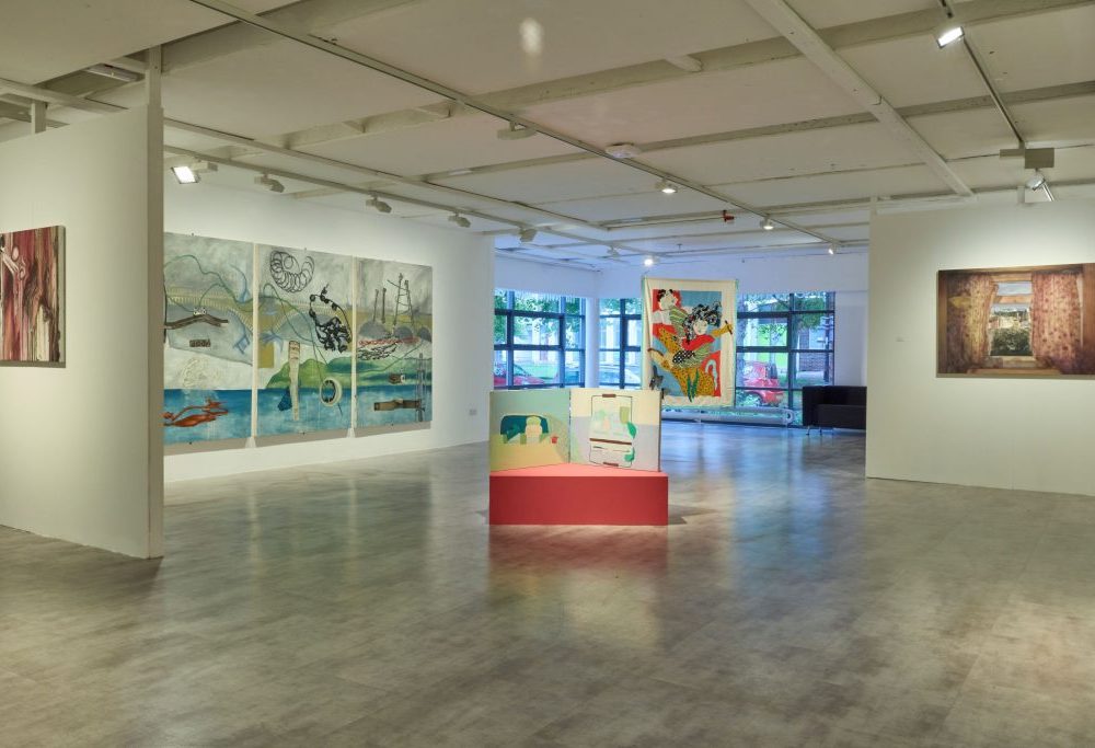 A bright gallery space with white walls displays large colorful paintings, including a multi-panel artwork and an abstract piece on a red pedestal in the center. Floor-to-ceiling windows in the background let in natural light.