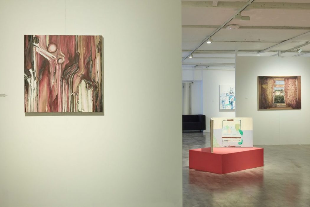 A close-up view of a gallery wall featuring an abstract painting in shades of red and brown. In the background, a red pedestal holds a small artwork, and other paintings are visible on adjacent walls.