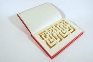 An open book with a red cover reveals intricate, layered paper cutouts forming a geometric maze carved into the pages, photographed against a white background.