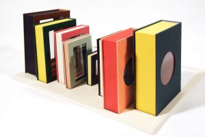 A row of sculptural artist books with brightly colored covers stands upright on a rectangular base, each featuring cut-out windows that reveal layered interiors. The books vary in size and color, creating a rhythmic, architectural arrangement against a white background.