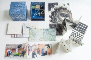 An arrangement of artist books and sculptural book objects is spread on a white surface, including folded accordion books, boxed pieces, and cut-paper constructions with photographic and abstract imagery. The works vary in shape and texture, featuring patterns, collaged scenes, and three-dimensional forms.