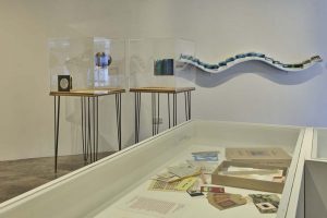A gallery display features tables with transparent cases containing sculptural book artworks, alongside a curved wall-mounted installation made of blue-toned pieces. A glass-topped display case in the foreground holds various printed materials and small objects.