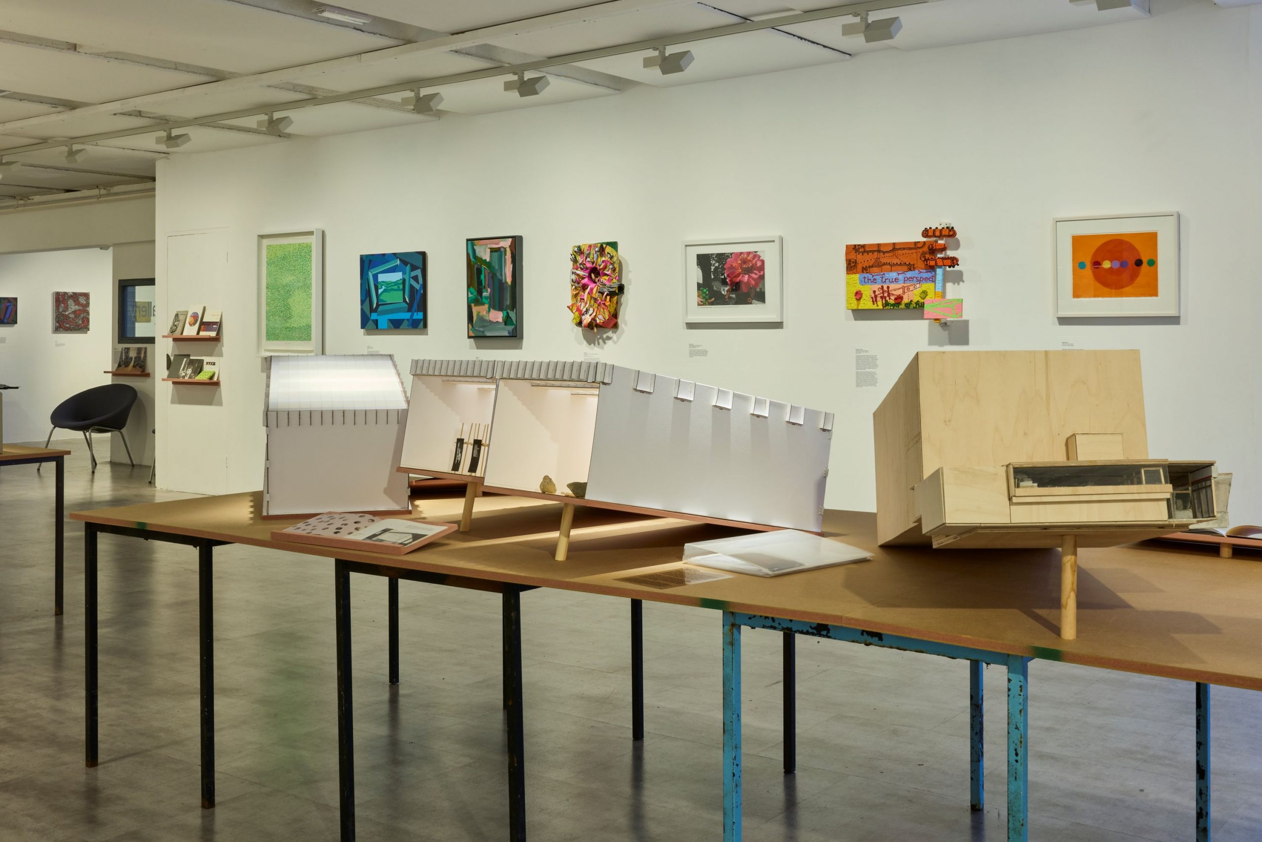 A row of architectural models is displayed on long tables in a brightly lit gallery, with vibrant paintings and prints mounted on the wall behind them. The models vary in shape and material, creating a visually layered exhibition.
