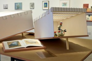 A detailed architectural model with an open side reveals a cluster of colourful spherical structures inside, displayed alongside an illustrated book on a table. Framed artworks hang on the wall behind the model in a gallery setting.
