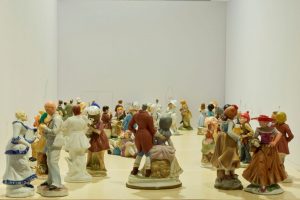 A large group of small ceramic figurines is arranged across a pale floor, each posed as if interacting or dancing. The figures fill the space from foreground to background within white gallery-style walls.