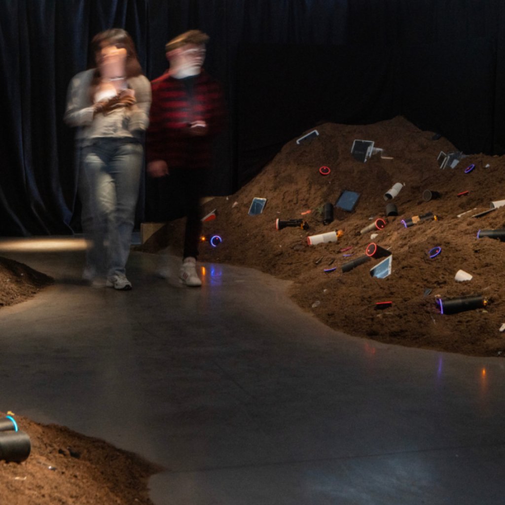 AI: Who’s Looking After Me? Exhibition featuring various modern electronic smart speaker devices placed randomly into dirt mounts. Two people are walking through the space.